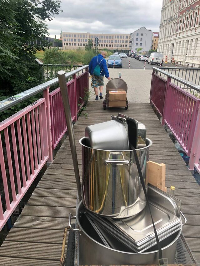Große Kochtöpfe und Gastrobehälter werden auf Wagen über eine Brücke transportiert.