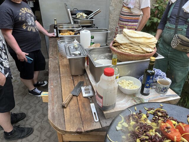 Ein langer Außentisch mit Fladenbrot, Gemüsepfanne, Küchenwerkzeug und Menschen beim Kochen.